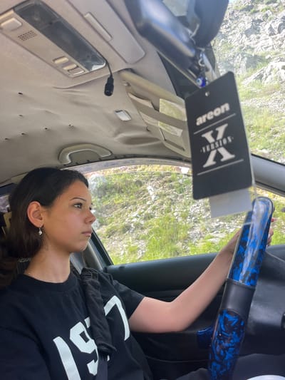 A young woman is driving a car with a blue patterned steering wheel. She is wearing a black t-shirt with white lettering that appears to say "19". A car freshener with the brand "areon" and "version X" is hanging from the rearview mirror. The car's interior is visible, including the headliner and a ceiling light. Outside the car, a rocky, grassy hillside is visible through the windows, suggesting a rural or mountainous setting. The location is identified as Vlahovo, Bulgaria.
