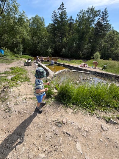 In Kul'dur, Russia, a young child wearing a bucket hat, striped t-shirt, and blue shorts stands on a dirt path, holding a stick. The child is facing a shallow, rocky pool of water bordered by stone. Several adults are seated or lounging by the pool, some in swimwear. The environment is lush and green, with trees forming a dense backdrop and tall grass growing along the water's edge. A wooden bench is visible to the left of the pool, and a blue tarp is partially seen further in the background. The scene appears to be an outdoor recreational area, possibly a natural hot spring or bathing spot, under a clear blue sky.