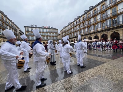 The image shows a group of people dressed in white chef uniforms and white hats, standing in a square in the city of Donostia, Spain (latitude: 43.324, longitude: -1.985). The square is paved with grey stone, and the surrounding buildings are old and have multiple stories.  The people are holding wooden sticks and appear to be drumming on barrels. There is a group of people dressed in colorful, traditional costumes standing in the background to the right of the group of chefs. They are also holding drums and instruments. The sky is overcast and the ground is wet.  The scene appears to be a street performance.