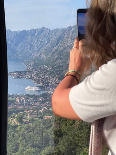 This image presents an elevated view overlooking a wide bay and a coastal town, identified as the area of Čavori, Montenegro, with rugged mountains forming the background. In the immediate foreground, the back and right side of a person are visible, facing away from the viewer towards the expansive landscape. The person is holding a light-colored smartphone with their right hand, appearing to capture a photo or video; the phone's screen reflects the distant scenery. A thin silver ring is present on the right ring finger, and two bracelets adorn the right wrist: one composed of light green and beige beads, and another black elastic band. The person wears a white, short-sleeved top with a ribbed texture, and brown hair falls over their right shoulder.
The midground and background reveal a large body of water, the Bay of Kotor, extending into the distance, bordered by a curving shoreline densely packed with urban development. Numerous buildings, predominantly residential, with varying roof colors, are scattered along the coast and climb the lower slopes. A large, white multi-deck cruise ship is anchored in the bay, positioned in the center-left. Several smaller boats are visible on the water, leaving faint wakes. The bay is framed by substantial, rocky mountains, partially covered in green vegetation, rising steeply from the water's edge. Below the immediate viewpoint, dense green trees cover the steep terrain, interspersed with individual buildings, including one with solar panels on its roof. The sky above is clear and light blue, indicating a sunny day.