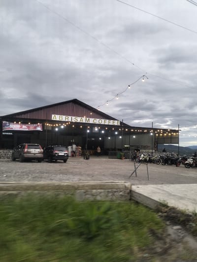 This media file captures an exterior view of Abrisam Coffee, a coffee shop in Panteraya, Indonesia. The scene depicts the building's facade with its prominent signage. The building has a dark, sloped roof and is illuminated by a string of lights running along its overhang. In front of the building, there's a gravel parking lot where several cars and numerous motorcycles are parked. A few people are visible, some near the entrance of the coffee shop and others near the motorcycles. The time of day appears to be late afternoon or early evening, indicated by the subdued natural light and the illuminated lights of the coffee shop. The sky is cloudy, suggesting overcast weather. The foreground shows a blurred patch of green grass, implying the photo might have been taken from a moving vehicle. Text visible includes "ABRISAM COFFEE" on the building's facade and "ABRISAM COFFEE NONTON BARENG" on a banner to the left.