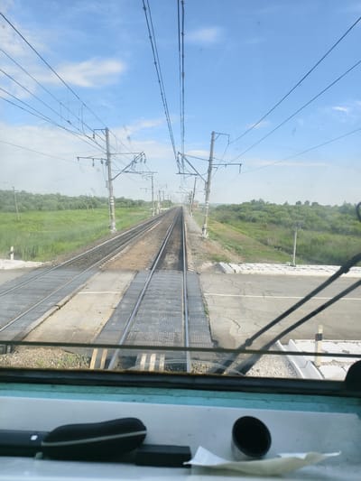 The image shows a view from inside a train cabin looking out at a railroad crossing. The sky is blue with some clouds, and the surrounding landscape is green with trees and grass. The railroad tracks extend into the distance, with power lines overhead. There is a road crossing the tracks, and some of the train cabin's interior is visible in the foreground, including a black handle and some white paper.

The image is likely taken in the city of Arkhara, Russia, a location known for its railway infrastructure. The scene appears peaceful and ordinary, depicting a typical view of a train traveling through a rural area. There are no people visible in the image, and no specific actions or interactions are occurring. The focus is on the environment and the infrastructure that supports train travel.