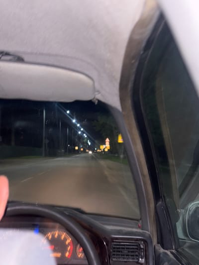 The image is taken from the interior of a car at night, looking out the windshield and side window. The driver's steering wheel and dashboard are visible in the foreground, with the speedometer illuminated. The road ahead is lined with streetlights and occasional signs, suggesting an urban or suburban environment. The scene depicts a car driving at night on a paved road. There is no information in the image that suggests the location is Ptichnik, Russia.