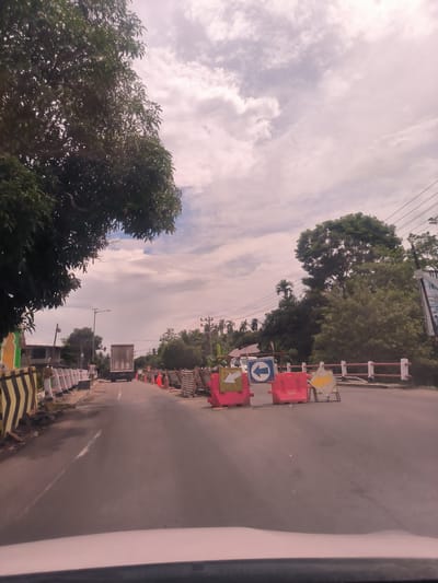 The image captures a daytime scene from the perspective of a vehicle driving along a road in Meukokuthang, Indonesia, facing forward. The sky above is overcast with scattered white and light grey clouds.

On the left side of the road, a large, lush green tree occupies the upper portion of the frame, its dense foliage extending downwards. Along the left edge of the road, yellow and black striped barriers are visible, followed by a series of white barriers lining what appears to be a sidewalk or raised median. Further down, a white truck is driving away from the viewer. Utility poles and power lines stretch across the sky in the mid-ground.

The right side of the road is heavily marked by construction or road work. Several bright pink plastic barriers are prominently displayed, along with orange traffic cones and wooden barricades. Amidst these, multiple road signs are visible: a blue circular sign with a white left-pointing arrow, a yellow square sign with a white right-pointing arrow, and a yellow diamond-shaped sign with a black arrow indicating a right and downward turn. Beyond the barriers, a dense array of green trees, including some palm trees, lines the road. A large sign or billboard, partially obscured and with unreadable text, stands on the far right.

The asphalt road surface features a faded white center line. The bottom of the image shows the dashboard of the vehicle, indicating the photo was taken from inside a car. The scene depicts an ordinary road with ongoing construction or maintenance activities.
