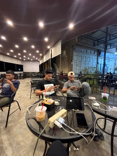 The image captures an evening scene at what appears to be an outdoor or semi-outdoor cafe or gathering space in Keudeublang, Indonesia. Three men are present, seated around black mesh-top tables. The lighting suggests it is nighttime, with numerous warm-toned lights embedded in a wooden or dark ceiling and a brightly lit building in the background.
In the foreground, a man wearing a dark blue t-shirt and olive green pants is seated, looking down at his phone. In the midground, another man in a black t-shirt is looking at his phone, with a white paper bag on the table next to him. To his right, a clear plastic cup with a red straw contains a light brown beverage, possibly iced coffee, with the word "HELLO" visible on it.
Further back, a third man wearing a grey polo shirt and a light-colored baseball cap sits with his legs bent, also focused on a device in his hands. He is seated next to a black mesh table holding various electronic devices and accessories. On this table, there is a black laptop, a dark smartphone, multiple white charging cables, and a beige power strip with "5" visible on its side. A white Samsung charger is plugged into the power strip. Another clear plastic cup with a green lid and red straw, containing a green beverage, is also on this table, along with a small white box, possibly a cigarette packet. Black sandals are visible under the table.
The background features a building with light blue lighting and distinct white paneled doors. Some text is faintly visible on the building in the distance. The overall atmosphere appears casual, with individuals engaged with their personal devices, possibly while socializing or relaxing.