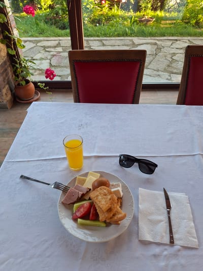 A breakfast table setting in Krumovgrad, Bulgaria. The table is covered with a white tablecloth and set with a plate of food consisting of eggs, cheese, ham, cucumbers, tomatoes, and toast, along with a fork, knife on a napkin, glass of orange juice, and sunglasses. Two chairs with red leather backs are positioned at the table, and a window with a view of green foliage and a stone exterior is visible behind the chairs. A potted plant with pink flowers sits on the windowsill.