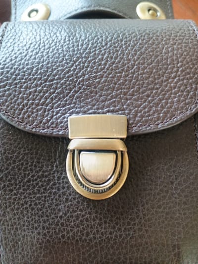 The image shows a close-up of a dark brown textured leather bag with a metal clasp. The clasp is silver-toned and has a decorative engraved edge. The leather has a pebbled texture, and there is visible stitching around the flap of the bag. The background is blurred, suggesting the focus is on the bag itself. There are no people or environmental details visible that would suggest a specific city or location. The image appears to be a product shot or a detail shot of the accessory.