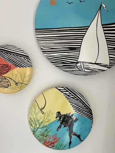 Three circular artworks are displayed on a white wall. The artwork at the top depicts a sailboat with two birds in the sky above a striped sea. The artwork on the left shows abstract shapes, including a coral-like element and seaweed. The artwork at the bottom features a scuba diver in the ocean, surrounded by coral and bubbles. All three artworks share a distinctive style with bold lines and vibrant colors. The image is taken from a close-up perspective, and no specific location in Ras Al-Khaimah, United Arab Emirates can be determined.
