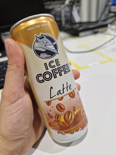 A hand holds a can of "Hell Ice Coffee Latte." The can is gold with a white and orange striped pattern. The logo at the top is a stylized devil's head. Text on the can reads "HELL ICE COFFEE" and "ICE COFFEE LATTE" in white and black lettering. The background is blurred and appears to be an indoor environment, possibly an office, with a white desk and some electronic equipment visible. The image does not contain any information related to the city of Bahovitsa, Bulgaria.