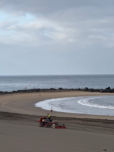 The image depicts a wide, sandy beach in Costa Adeje, Spain, under an overcast sky. In the foreground, a red and orange beach tractor with a grooming attachment is actively working on the sand, leaving parallel tracks. Two individuals are visible near the tractor, one standing and the other appearing to be operating the machinery. Further back, a person in an orange shirt stands on a dark, rocky breakwater, and another person in a white shirt stands nearby on the same structure. The ocean stretches to the horizon, with a white boat visible in the distance. Gentle waves are breaking on the shore.