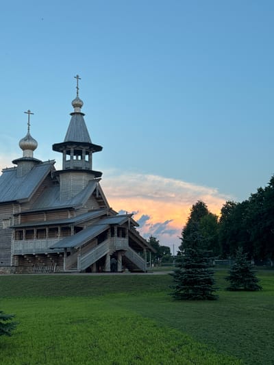An outdoor, medium shot captures a wooden, traditional architectural style structure with multiple levels and tiered roofing, topped with onion domes and crosses, set against a twilight sky with wispy clouds tinged with orange and pink. The building's exterior is constructed from weathered wooden logs and features intricate wooden carvings and latticework on its balconies and staircases. Two pine trees stand in the foreground on a manicured green lawn, with a line of darker trees forming the background to the right. The sky is a gradient of light blue to a softer hue near the horizon, indicating early evening. No people are visible in the frame. The overall scene suggests a historical or cultural site, possibly a church or museum, in the Нёманскі сельскі Савет, Belarus.