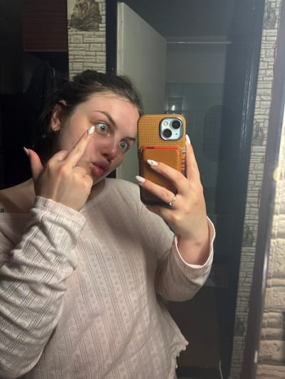A woman is taking a selfie in what appears to be a mirror. She is holding a phone with a brown case and is pointing at her eye with her left hand. She is wearing a light pink, long-sleeved, patterned shirt. Her hair is dark and pulled back. The background shows a room with tiled walls and a dark-colored shelf or ledge. The location cannot be determined, including the city of Chornomorskoe, Ukraine.