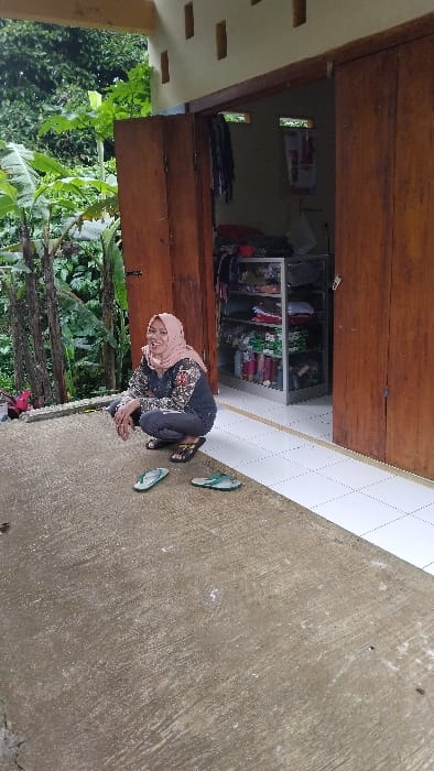 The image depicts a woman crouching on a concrete surface outside a building, set in a semi-urban or rural environment in Kecamatan Panjalu, Indonesia.

**Subjects:** The primary subject is a woman, wearing a light-colored hijab, a patterned long-sleeved top, and dark trousers. She is in a crouched position, looking towards the viewer with a slight smile.

**Objects:** Two pairs of flip-flops are visible on the concrete ground near the woman – one dark pair and one light green pair. Inside the building, through an open doorway, a white metal shelving unit is visible, stocked with various items, likely merchandise such as clothing or packaged goods, suggesting a small shop or storage area.

**Scene and Setting:** The scene is set outdoors on a rough concrete surface. To the left, a dense growth of green foliage and trees indicates a natural, possibly overgrown, area. The building features wooden elements around its entrance. Inside, a white tiled floor contrasts with the darker interior. A light-colored wall or pillar with several rectangular cutouts is present on the far left.

**Activity or Event:** The woman is casually crouching, possibly resting or posing for the photograph, engaging directly with the camera through her gaze and smile.

**Notable Details:** The image is illuminated by natural daylight, indicating it is daytime. The overall atmosphere appears calm and unhurried. The simple concrete ground, wooden and tiled building elements, and surrounding greenery are consistent with a local Indonesian setting. No discernible text is visible in the image.