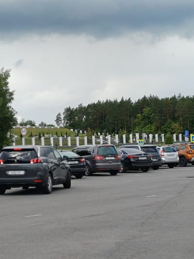 A line of cars are parked or stopped on a paved surface in front of a landscaped area with white posts and trees. The sky is overcast. A speed limit sign showing "60" is visible on the left. License plates on the cars suggest they are from Belarus.