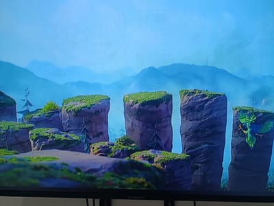 The image shows a digital rendering of a mountainous landscape with towering, moss-covered rock formations. The scene is bathed in soft, ethereal light, suggesting either dawn or dusk. In the distance, layers of misty mountains recede into a hazy blue sky. The foreground is dotted with patches of green grass and smaller rock clusters, creating a sense of depth and scale. A stylized, bird-like creature with vibrant green and blue plumage is perched on the side of one of the large rock pillars on the right. The overall atmosphere is serene and fantastical, with no visible people or explicit indication of a specific event. The word "SKYWORTH" is visible in white text at the bottom of the screen, indicating this is likely a screenshot from a television or display. The location context provided, Arkadag, Turkmenistan, does not appear to be directly depicted in the imagery itself, but rather serves as a contextual marker for the origin of the media.
