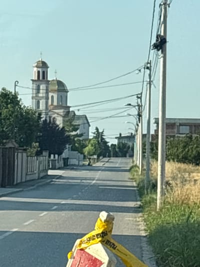 This image captures a street scene in Surcin, Serbia, possibly on a bright, sunny day given the clear blue sky and strong shadows. The photograph is oriented vertically.
The primary subjects include a long road with a lane marked by white lines, flanked by buildings and vegetation. In the foreground, a yellow barrier tape, partially obscured by glare, is visible on the right side of the road, suggesting potential construction or a restricted area. The tape has visible text, though it is difficult to fully discern due to the angle and resolution; some letters appear to read "POLICIJA" (Police in Serbian) and possibly "STOP".
Further down the road, a prominent building with multiple domes and architectural features resembling a church or religious institution is visible on the left. The building has a white facade, and the domes are a muted green or gold color. Trees and other greenery surround this structure and line the street. Utility poles with numerous wires stretch across the upper portion of the image, indicating the presence of power lines and possibly communication infrastructure.
The overall scene depicts a typical day on a road in a Serbian urban or semi-urban area, with a mix of modern infrastructure, religious architecture, and natural elements. The time of day appears to be during daylight hours, likely morning or afternoon, due to the lighting and the appearance of the sky. No people are clearly visible in the frame.