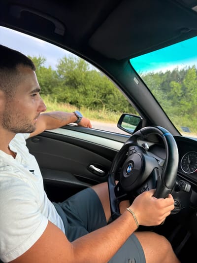 A young man with short dark hair and stubble is driving a car, seen from the passenger's perspective. He is wearing a white t-shirt and dark shorts. His left arm is resting on the open car door, and he is looking out the window. On his wrist, he is wearing a smartwatch. The car's interior is visible, including the steering wheel, dashboard, and a side mirror. Outside the car, lush green trees and vegetation are visible. The sky appears to be a bright, clear blue. The scene appears to be a rural setting, possibly on a road outside of Хаджийско, Bulgaria.