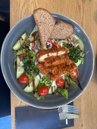 A top-down image depicts a meal served on a light-colored wooden table in Weißenburg in Bayern, Germany. The primary subject is a large, circular, matte gray-blue ceramic bowl filled with a salad. The salad comprises various leafy greens, semi-circular cucumber slices, whole and halved red cherry tomatoes, thin red onion rings, and small groupings of dark green foliage. A visible creamy white dressing coats the salad components, augmented by a dark, viscous drizzle, consistent with balsamic glaze. Centrally placed are five segments of golden-brown, breaded chicken, one of which is bisected, exposing white poultry meat. Two slices of dark, dense-grained bread, one upright and one angled, are positioned at the upper edge of the bowl. Adjacent to the lower right of the bowl, a stainless steel fork and knife are partially visible, secured within a black paper wrap or napkin. The immediate environment consists of a light-toned wooden table surface with discernible grain, extending beneath the bowl and cutlery. A blurred dark object occupies the far left background.