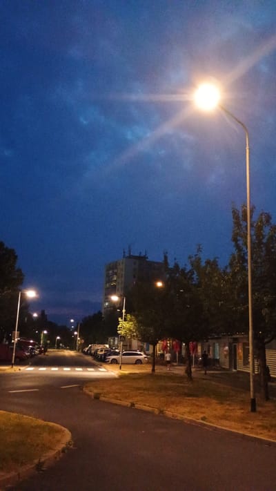This image depicts a street scene at dusk in Sokolov, Czechia. The sky is a deep blue, with a bright streetlamp in the upper left corner casting a strong glare. In the mid-ground, a tall, multi-story building stands out against the darkening sky, with its windows lit. Trees line the street, some of which are illuminated by streetlights. To the right of the building, a road curves away, bordered by a grassy verge. Several cars are parked along the side of the road, and a few figures, likely pedestrians, can be seen walking in the distance. The overall atmosphere suggests a quiet evening, with the artificial lights beginning to dominate the natural light as day transitions into night. The weather appears clear. No visible text is present in the image.