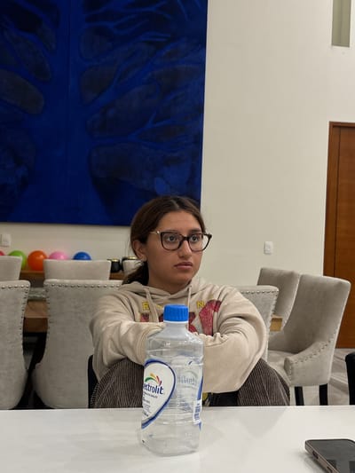 The image shows a woman with brown hair and glasses sitting at a table with a bottle of Electrolit water. The background includes a blue painting, chairs, and balloons. The location is likely Santiago Etla, Mexico.