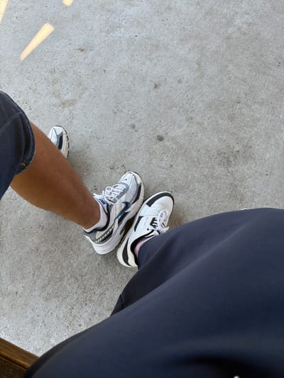 The image shows two pairs of feet wearing athletic shoes on a concrete surface. One person is wearing dark shorts and white and blue Nike sneakers. The other person is wearing dark pants and white Skechers sneakers. The concrete surface appears to be outdoors, possibly a sidewalk or patio. There are some shadows cast on the concrete from an unseen light source, suggesting it is daytime. The scene does not provide any specific details or context that would place it in Dinkata, Bulgaria.