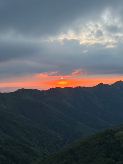 The image captures a panoramic view of a mountainous landscape, likely near the city of Rozino, Bulgaria, during a sunset. The foreground is dominated by the dark, densely forested slopes of multiple mountain ridges, appearing as layers receding into the distance. The trees are a deep, muted green, suggesting either the shadows of the setting sun or the natural coloring of the foliage.
The horizon is marked by a fiery band of orange and red, indicative of a dramatic sunset. The sun itself is a bright, almost white orb, partially obscured by the atmosphere, positioned on the left side of the horizon. Above the sunset colors, the sky transitions to a soft pink and then to a dusky blue-gray. Further up, the sky is filled with heavy, dark gray clouds that are breaking in places to reveal hints of lighter, brighter sky, possibly where the sun's last rays are filtering through.
The mountains are rugged and textured, with visible contours and undulations. The scale suggests a vast, natural expanse. There are no people, animals, or man-made structures clearly visible in the image, emphasizing the wild and untouched nature of the environment. The overall mood is serene and awe-inspiring, highlighting the beauty of a natural sunset over a mountainous terrain.