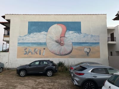 The image captures a mural painted on the exterior wall of a building in Toroni Municipal Unit, Greece. The mural depicts a beach scene with a large, stylized nautilus shell in the center, surrounded by clouds and the sea. The word "SARTI" is painted in large, stone-like letters on the sand. To the right of the nautilus shell, a heart shape formed by two stones is depicted. In the foreground, three cars are parked on a dirt surface. The building itself appears to be a multi-story structure with balconies. The sky is overcast, suggesting it is either early morning or late evening. The overall scene is a colorful and artistic representation of a coastal landscape.