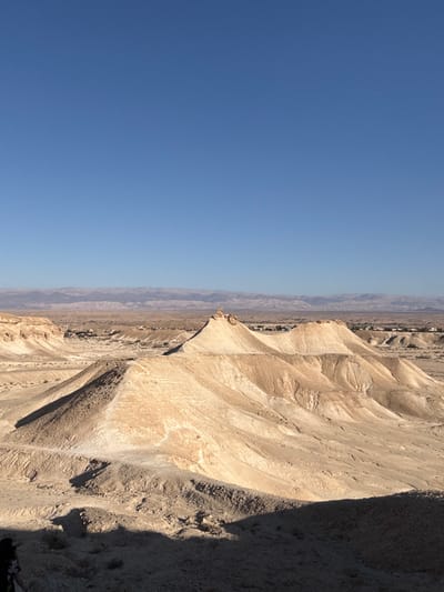 This image captures a wide-angle desert landscape within the Arava Tichona Regional Council, Israel, under a clear, cloudless blue sky that occupies the upper third of the frame. The foreground, at the bottom left, is heavily shadowed, partially revealing the head and upper torso of an individual, likely wearing a dark hat or head covering, observing the scene. The mid-ground is dominated by a series of undulating, light tan and off-white eroded hills and sand formations, characterized by deep contours and sharp shadows. Several of these formations exhibit distinct, somewhat conical or dome-shaped peaks with darker, weathered caps. In the distant mid-ground, nestled within a valley between the hills and further mountain ranges, a small settlement or town is visible, consisting of scattered light-colored buildings and minimal green vegetation. The far background presents a hazy, layered range of muted grey-brown mountains stretching horizontally across the horizon. The scene is illuminated by strong, direct sunlight, casting prominent shadows that define the geological features.