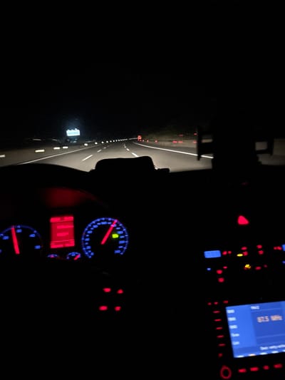 The image depicts the interior of a car driving on a highway at night. The viewpoint is from the driver's seat, looking forward. The dashboard instruments are illuminated, with a speedometer showing approximately 120 km/h and a tachometer showing around 2000 RPM. The car's radio display is also visible, showing "87.5 MHz". The highway ahead is lit by streetlights and the headlights of other vehicles, creating streaks of light. The background is dark, indicating nighttime. Based on contextual information, the location is identified as the city of Ergene, Turkey. There are no people clearly visible in the frame, only the implied presence of a driver.