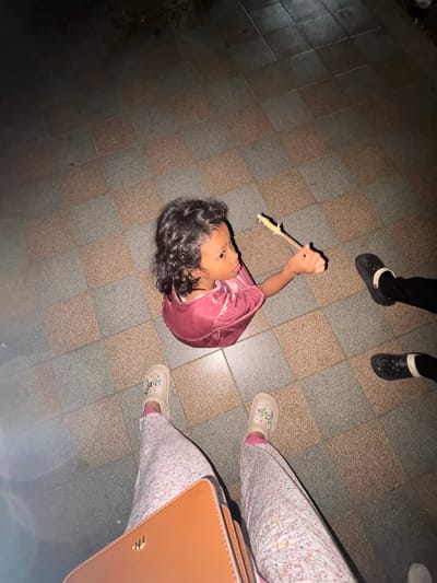 In Tanggul, Indonesia, a child with curly dark hair, wearing a pink dress, stands on a tiled ground. The child holds a light-colored object on a stick. Two individuals, whose legs are visible, are also present. One wears black Crocs and black pants, while the other wears light-colored clogs with floral designs and floral-patterned pants. A brown purse is held by the person in floral pants. The setting appears to be outdoors, possibly a pathway or courtyard, with low lighting.
