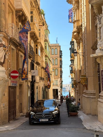 The image depicts a street scene in Valletta, Malta, characterized by its historic architecture and narrow passageways. The buildings, constructed from a warm-toned stone, feature ornate balconies and intricate facades. Several flags with a royal emblem and depicted figures are hung across the street, adding a splash of color and cultural significance.
A black Mercedes-Benz car is parked on the right side of the street, its license plate reading "DLY-927". The car's reflection in the building opposite suggests a sunny day. In the background, more buildings rise up, some painted in vibrant green, and a glimpse of the sea and a distant cityscape is visible, hinting at the city's coastal location.
Several people are scattered throughout the scene. Some are seated at outdoor tables in front of a building, possibly a cafe or restaurant, enjoying the day. Others are walking along the street. The overall atmosphere appears relaxed and leisurely. A circular sign with a red border and a white bar inside is mounted on a wall, possibly indicating a restriction or prohibition. Further down, a sign reads "FAIR GAME STORE..", suggesting a retail establishment. The presence of decorative lighting strung across the buildings and the clear blue sky indicate it is daytime. The architecture and the style of the flags strongly suggest a Mediterranean setting, consistent with Valletta, Malta.