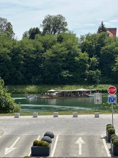 The image shows the Drava River in Maribor, Slovenia. A floating platform with people is visible on the river. A stop sign and a blue sign with a bicycle and pedestrian symbol and the number 20 are near the river. A brick walkway with a white arrow is on the right side of the image, with plants in containers lining the walkway. Trees cover the riverbank and extend into the background.