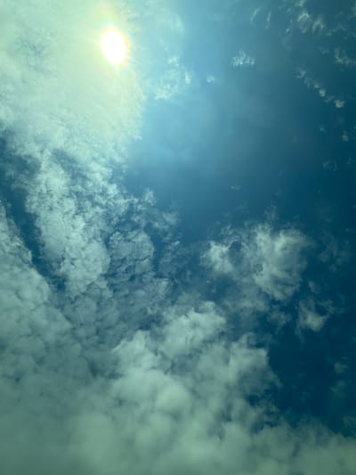 A low-angle shot depicts a bright sun positioned in the lower-left corner, surrounded by thin, layered clouds. The sky transitions from a clear, bright blue in the upper-left to a dense cloud cover occupying the majority of the frame. The clouds are predominantly white, with some appearing slightly gray. The environment suggests an outdoor setting with natural light. There are no discernible people, subjects, objects, actions, or interactions in the image, apart from the sky itself and its natural elements. The image was taken in Manolsko Konare, Bulgaria.