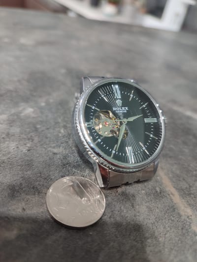An overhead, eye-level shot shows a silver wristwatch and a coin positioned on a dark, textured surface that resembles concrete. The watch is on the right side of the frame, with its black face displaying Roman numerals and a visible inner mechanism. The watchband is also silver. To the left of the watch, a silver coin lies flat on the surface. The background is blurred, suggesting an indoor setting with diffused lighting. There is no indication of the city of Grigoriopol, Moldova, or any specific details about the environment that would place it there.