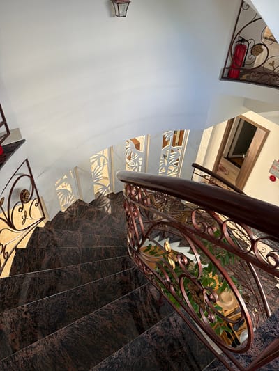 The image shows a downward view of a spiral staircase with dark, speckled granite steps and a dark red metal railing featuring ornate scrollwork and floral motifs. The walls are painted white, and the ceiling is also white, with a rustic lantern fixture mounted on the upper wall. To the right, on an upper landing, a red fire extinguisher is visible next to a decorative metal gate. Further down the staircase, there are patterned white screens with a tropical leaf design on the wall. On a lower landing, a doorway is open, revealing a glimpse of an interior space with wooden furniture and some items, including what appears to be a hat. The lighting suggests it is daytime, with sunlight casting shadows on the stairs and illuminating the decorative elements. The scene is an interior staircase, likely in a residential or commercial building, located in Cam Lâm, Vietnam, as per the provided context.