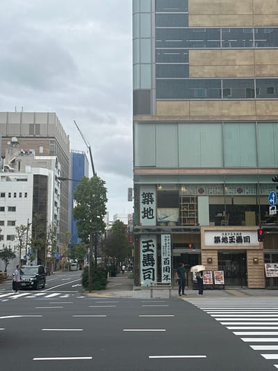 The image depicts an urban street scene in Chūō, Japan, specifically the Tsukiji district, as evidenced by prominent "築地" (Tsukiji) and "築地玉寿司" (Tsukiji Tamasushi) signage. The foreground features a dark asphalt road with white dashed lane markings and a white zebra crosswalk on the right. A black Nissan taxi is visible in the left lane, moving from the background towards the foreground. A male pedestrian in a plaid shirt walks on the left sidewalk, observing the approaching taxi.

On the right side of the image, a multi-story commercial building houses the "Tsukiji Tamasushi" restaurant on its ground floor. The restaurant exhibits a light wooden facade with menu displays and features two large vertical signs in Japanese script, one prominently displaying "築地" and the other "玉寿司" with additional text. A red traffic light is positioned above the restaurant entrance. Two individuals are present on the sidewalk in front of the restaurant: a male in a dark green t-shirt and blue jeans stands with his hands in his pockets, and to his right, another individual holds a white umbrella over their head, dressed in dark clothing. Both individuals face generally across the street or towards the intersection.

The background on the left reveals a street lined with various multi-story urban buildings, predominantly light-colored. A construction crane is visible above these buildings in the distance, and a building under construction is partially covered in blue scaffolding. The upper floors of the building on the right feature a facade composed of alternating horizontal bands of light beige panels and dark, reflective glass windows. The sky is uniformly overcast.