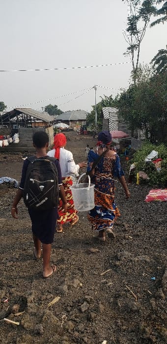 The image depicts three individuals walking along an unpaved, dark brown dirt path in Buzove, Democratic Republic of the Congo. The foremost person, wearing a dark garment and light-colored flip-flops, carries a black backpack with white markings. Behind them, a second individual wears a white long-sleeved shirt, a red headscarf, and a long, patterned skirt in red, orange, and yellow. The third person, to the right, wears a blue and orange patterned dress, a blue head wrap, and carries a transparent plastic bucket containing unidentifiable dark objects in their right hand. All three individuals are walking away from the camera.

The immediate environment consists of loose soil and gravel. In the background, various single-story structures with corrugated metal or dark roofs are visible. On the left, an open-sided building with an elevated platform and stairs is present, with white laundry hanging from its railings. On the right, dense green foliage and discarded items are visible near the path. A utility pole with overhead lines extends into the scene. The sky is overcast and light grey.