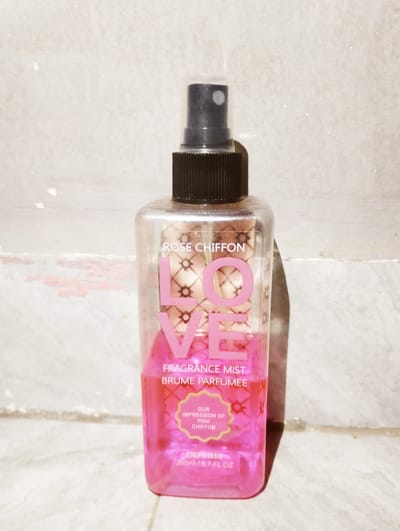 The image shows a bottle of "LOVE Rose Chiffon" fragrance mist. The bottle is clear and square-shaped, filled with a pink liquid. The label features large pink lettering spelling out "LOVE" vertically. Above it, the text "ROSE CHIFFON" is visible, and below "LOVE," it reads "FRAGRANCE MIST BRUME PARFUMEE." A smaller section of the label contains text within a yellow, scalloped-edged shape, mentioning "OUR IMPRESSION OF PINK CHIFFON." At the bottom, "DIPERIO 200ml / 6.7 FL OZ" is printed. The bottle is placed in front of a textured, light-colored background that appears to be a wall or textured surface. The lighting suggests it might be daytime, with shadows cast to the right of the bottle. The image appears to be taken indoors or in a sheltered outdoor space, given the static background. There are no people or animals in the image. The setting does not provide specific location cues beyond it being a visually plain background suitable for product photography. The overall impression is that of a product shot for a cosmetic or personal care item.