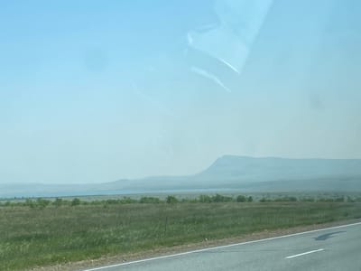 This is a horizontal, medium-shot photograph taken from a vehicle, looking out over a vast, open landscape under a hazy, pale blue sky. The foreground shows a paved road with a white line marking its edge. Beyond the road is a wide expanse of green grassland, with some scattered shrubs and small trees. In the middle ground, a body of water, possibly a lake or a wide river, is visible, bordered by a line of trees. The background is dominated by rolling hills and a prominent, flat-topped mountain, all appearing muted and indistinct due to atmospheric haze. The scene suggests a rural or natural environment, possibly in a steppe or prairie region. The image does not depict the city of Aeshka, Russia, nor does it contain any people or specific actions.