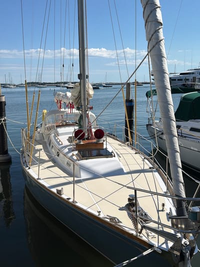 The image shows a sailboat docked in the Village of Sag Harbor, United States of America. The sailboat is the primary subject, featuring a white hull with a blue stripe along the waterline, a beige deck, and a tall mast. Several lines and sails are visible, some furled and tied. The boat is moored next to a dock, with pilings and ropes securing it. In the background, other boats and a large white yacht are visible on the water under a clear blue sky with scattered clouds. The overall scene depicts a peaceful harbor setting on a sunny day. There are no people visible in the image.