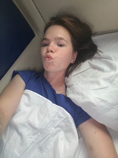 A young woman with auburn hair and freckles is lying on a bed, looking at the camera and puckering her lips. She is wearing a blue t-shirt and is partially covered by a white sheet. The room appears to be dimly lit. The background shows a blue wall and a beige ceiling.