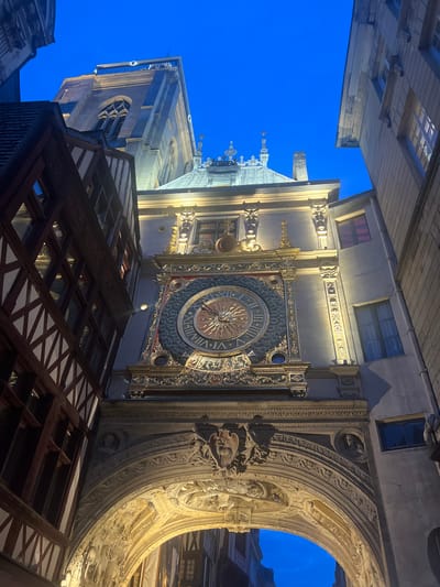 The image presents a low-angle view of the Gros Horloge (Great Clock) and its archway in Rouen, France, during nighttime or late twilight.

At the center, the ornate Renaissance-style archway of the Gros Horloge is prominently illuminated by warm artificial lighting. The archway features elaborate carved bas-reliefs depicting figures and scenes, and medallions containing sculpted busts are visible on the spandrels. Faint inscriptions are present on the inner curve of the arch. Above the archway, the astronomical clock face is visible. It is a large, circular dial with a dark, textured background, ornate gilded borders, and a central sunburst motif. Roman numerals from VI to III are clearly visible, and a single gilded hand points to approximately 10:10. Above and around the clock face, the structure is highly decorated with gilded pilasters, pediments, and sculptural elements, all brightly lit.

To the left of the clock structure, a traditional half-timbered building with dark wooden beams and light-colored infill rises vertically. Its windows are dark. To the right, a multi-story stone or stucco building with several rectangular windows, some displaying reddish or bluish tints, frames the clock.

Above the clock and to the upper left, a grey stone church tower with a tall, pointed Gothic arched window and a castellated top is visible against the deep blue sky. Other rooflines and structural elements are faintly visible against the sky in the upper right. The artificial lighting accentuates the architectural details of the clock and archway, while the surrounding buildings and the sky recede into the dim light. No people are present in the scene.