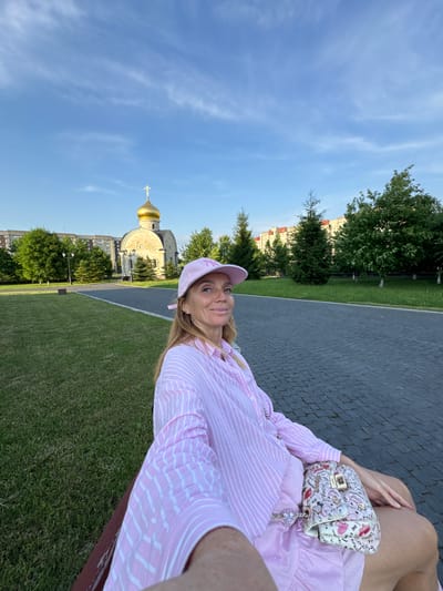 A woman wearing a pink striped shirt and a pink baseball cap sits on a park bench in Kirishi, Russia. A small, ornate church with a golden dome and cross is visible behind her, with residential buildings and trees in the background. The woman is holding a purse adorned with a pattern of lips and text. The ground is paved with cobblestones and a grassy lawn is to her left. The sky is blue with scattered white clouds.