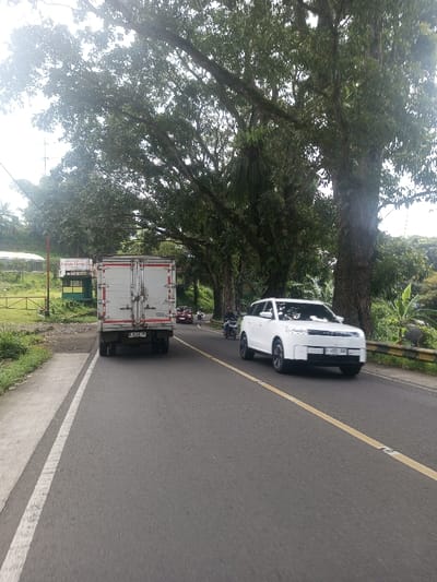 The image depicts a two-lane paved road in Niz Lakuk, India, under an overcast sky. A white box truck with license plate "B 8359 FP" is positioned in the left lane, traveling away from the viewer. Its rear doors are visible. Adjacent to the road's left side, behind the truck, is a grassy area featuring a green structure with a partial sign reading "Langit C..." and a reddish pole. A white SUV, license plate "B 4569 OU", occupies the right lane, moving towards the viewer. Further down the road, behind the SUV, multiple motorcycles and at least one red car are visible, also moving away from the viewer. The road is marked with a solid white line on the left shoulder and a dashed yellow center line. Large, mature trees with dense green foliage line both sides of the road, forming a canopy overhead. The trees on the right exhibit visible moss or epiphytes on their trunks and branches. A yellow and black striped guardrail is present along the right edge of the road, with additional dense vegetation, including what appear to be banana plants, beyond it. No individuals are clearly visible.