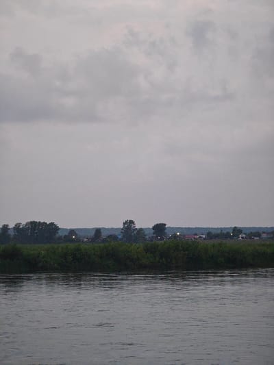 The image depicts a tranquil, overcast scene at dusk or dawn on the Angara River in Kazachinskoye, Russia. The foreground is dominated by the calm, rippling surface of the river, reflecting the muted tones of the sky. Beyond the river, a dense line of green foliage forms a natural barrier. In the middle ground, a distant settlement is visible, characterized by scattered houses with illuminated windows, suggesting human habitation. The background is filled with a hazy, grey sky, with soft, indistinct clouds. The overall atmosphere is one of quietude and stillness, with no visible people or active events. The image focuses on the natural landscape and the subtle signs of a remote community.