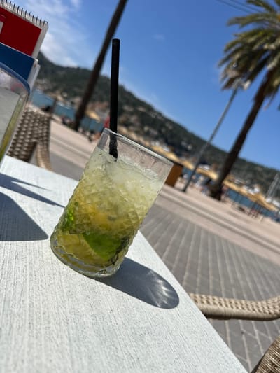 The image shows a close-up of a cocktail glass filled with a light-colored drink, presumably a mojito, due to the presence of lime and ice. A black straw is inserted into the drink. The glass sits on a white, textured table. Behind the table, the blurred background reveals a coastal scene with palm trees, a beach, and water. The image seems to be taken outdoors, likely in El Port, Spain. There are no people directly visible in the frame, but there are other objects in the table that are not easy to see. The overall scene suggests a relaxed, vacation atmosphere.