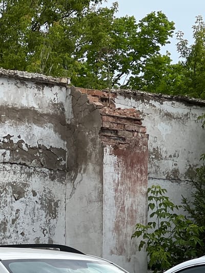 The image shows a damaged exterior wall of a building. The wall is made of brick and plaster, with significant sections of plaster missing, revealing the brickwork underneath. The top edge of the wall is also damaged, with bricks exposed and crumbling. Lush green trees are visible behind and to the side of the wall. The lower part of the image shows the roof and windshield of a white car. The scene appears to be an outdoor environment, likely in a city given the presence of a car and a building. There are no people visible in the image. The overall impression is one of disrepair and neglect.