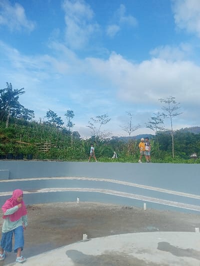 This image captures an outdoor scene, likely a public or recreational area in Pangalengan, Indonesia, featuring a terraced concrete structure with people. The foreground shows the lower levels of a grey and white concrete amphitheater or terraced seating area, with several white pipes protruding from the ground.
In the bottom left, a young girl, wearing a pink hijab, a light green long-sleeved shirt, blue pants, and sandals, walks leftward, looking down. Her shadow is visible on the concrete.
On the upper level of the terraced structure, three young boys or teenagers are present. One boy, dressed in a light-colored shirt and shorts, walks from left to right. Further to the right, two other boys stand close together, seemingly conversing. One wears a yellow patterned shirt and shorts, while the other has a light blue shirt, shorts, and a cap.
Behind the concrete structure, a vibrant green hillside rises, densely covered with trees and various vegetation, including what appear to be agricultural plantings in black polybags and some bamboo-like trellises. Sparse, thin-branched trees dot the landscape. Distant mountains or hills are visible in the far background.
The sky is bright blue with scattered white, fluffy clouds, indicating clear and pleasant weather, likely during the morning or early afternoon. The scene conveys a relaxed outdoor setting, possibly a local community space or a scenic viewpoint. There is no visible text in the image.