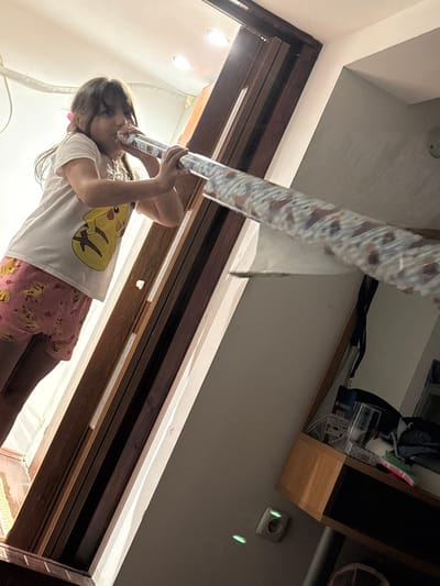 A young girl, wearing a white t-shirt with a Pikachu graphic and pink shorts with a similar pattern, stands indoors, holding a long, rolled-up object to her mouth as if speaking into it. The object appears to be a tube wrapped in patterned paper. The background suggests an indoor environment with wooden door frames and shelves. The lighting is dim, with some overhead light sources visible. The scene is likely a residential setting in Enevo, Bulgaria.