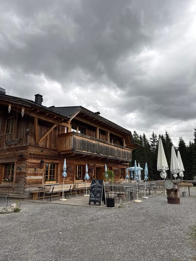 An image captures a rustic, multi-story wooden building, likely an alpine hut or restaurant, set against a backdrop of dense evergreen trees under an overcast sky in Untergiblen, Austria. The building, constructed from dark, weathered timber planks, features multiple windows and a balcony on its upper level.

In front of the building, a large gravel patio serves as an outdoor seating area, furnished with numerous tables and chairs. Several large white and light blue parasols, some partially closed, provide potential shade for diners. No people are visible, suggesting the area is currently unoccupied.

Near the building, two blackboards display menu items, including "HUGO" for €7.50, "APEROL," "HOT APEROL," and "ALKOHOLF[REI]" (alcohol-free), along with other numbers like "€3,50" and "0." A sign affixed to the building clearly reads "KASERMANDL SPECKHOLLE."

The sky is filled with thick, grey clouds, indicating overcast weather and diffused daylight. The ground is covered entirely in light grey gravel. In the background, beyond the patio, tall evergreen trees rise towards the cloudy sky. A large, rusted metal barrel or container stands at the far end of the seating area.